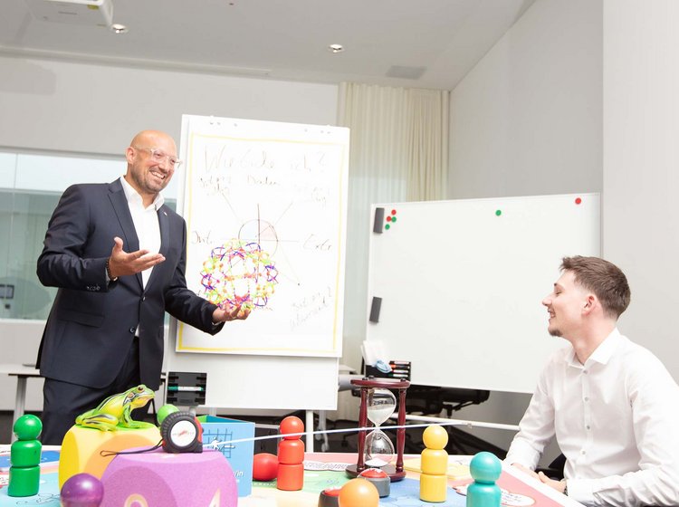 Sales seminar A man in a suit is giving a presentation to two seated colleagues, using a flipchart with colorful drawings, while they listen and engage attentively in a bright meeting room.