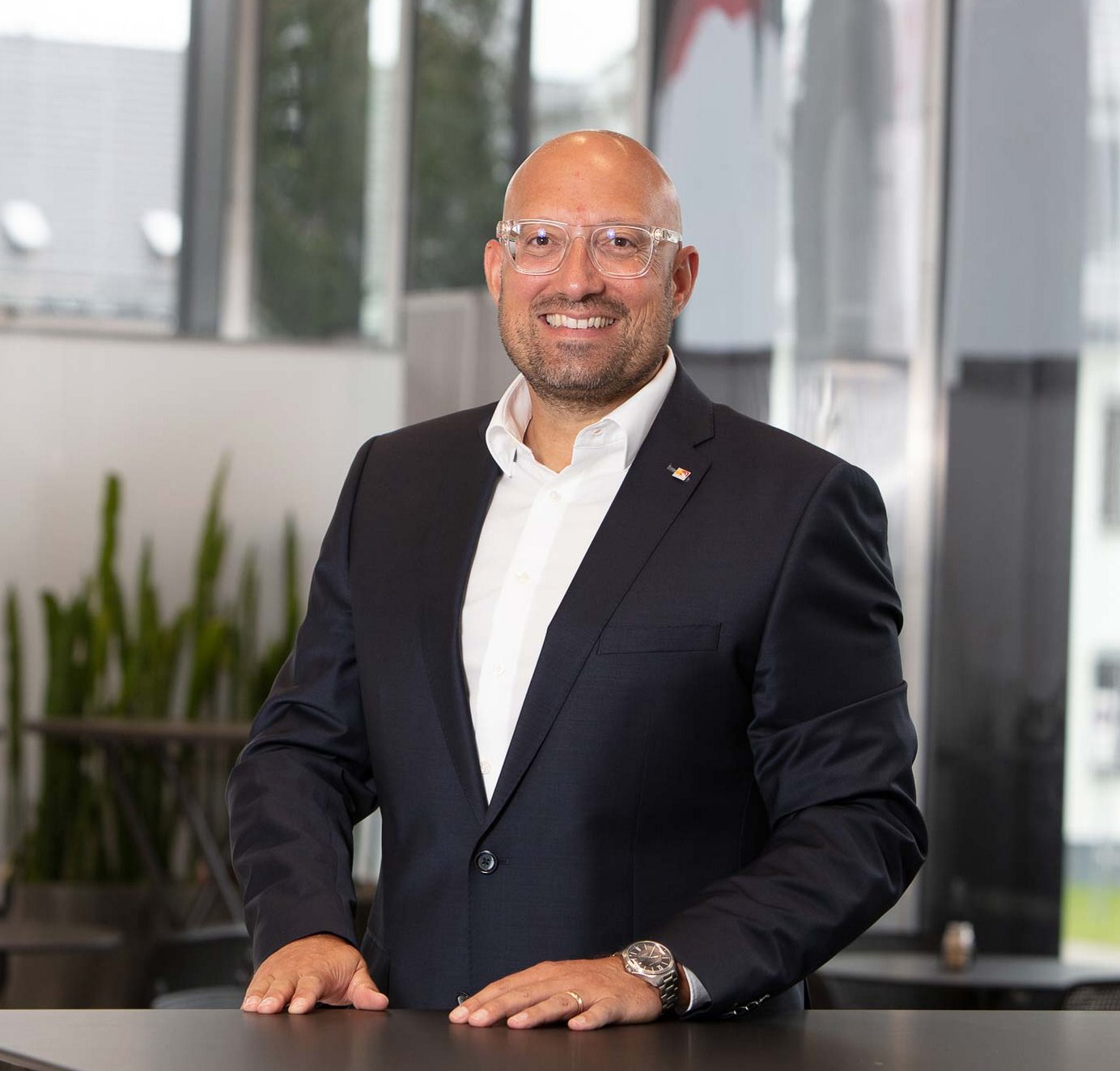 Sales training by Philipp Gotterbarm A confident man wearing a dark suit and white shirt stands smiling behind a table in a modern indoor space with large windows.