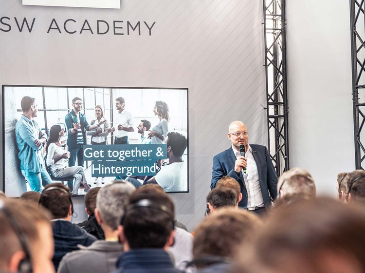 Presentation by Philipp Gotterbarm Ein Mann hält eine Präsentation vor einem Publikum in einem Seminarraum der PSW Academy. Auf einem Bildschirm im Hintergrund ist eine Gruppe von Menschen zu sehen, darunter der Text „Get together & Hintergrund“.