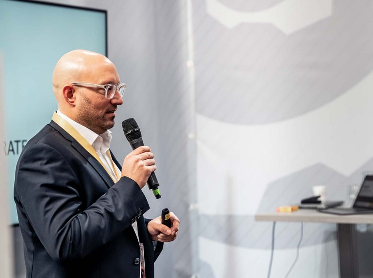 Sales consultation with Philipp Gotterbarm A man in a suit is speaking into a microphone, delivering a presentation or speech in an indoor setting.