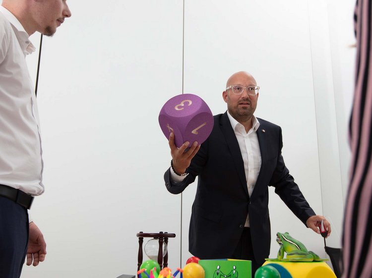 Sales training with Ludoki Two men participate in an interactive activity involving colorful oversized objects, including a large purple die and various shapes, on a table in a minimalistic room.