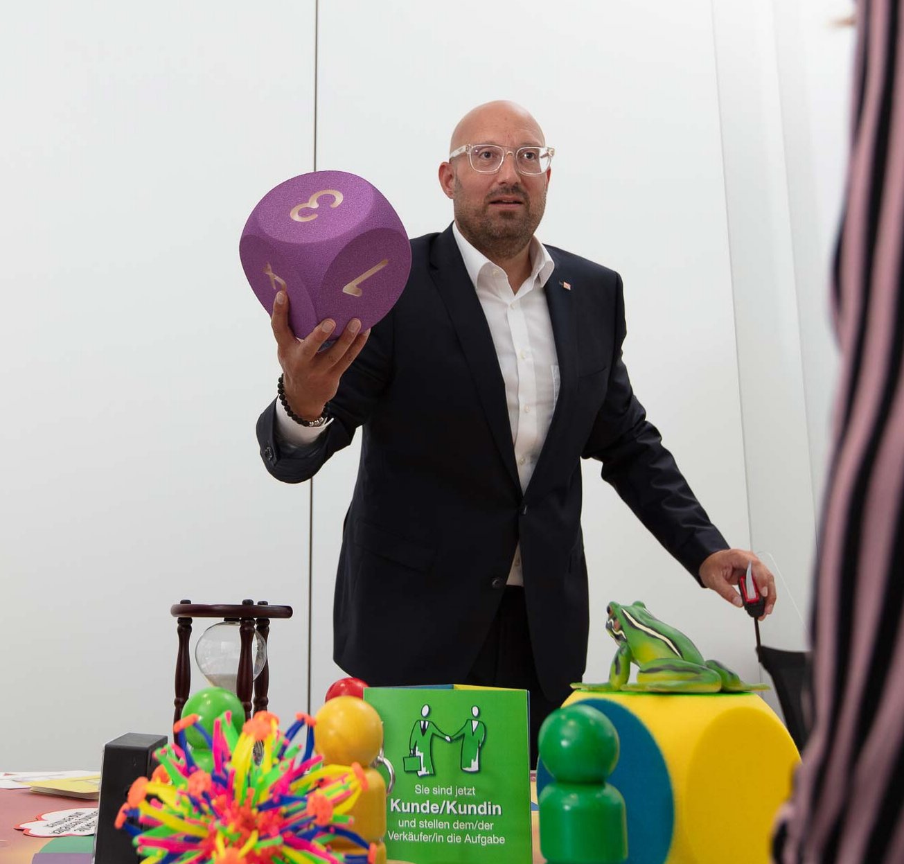 Sales training with Ludoki Two men participate in an interactive activity involving colorful oversized objects, including a large purple die and various shapes, on a table in a minimalistic room.