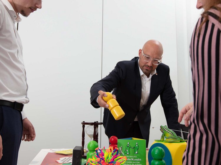 Sales training with Philipp Gotterbarm Three people are engaged in a collaborative activity, interacting with colorful educational tools and objects on a table in a white room.