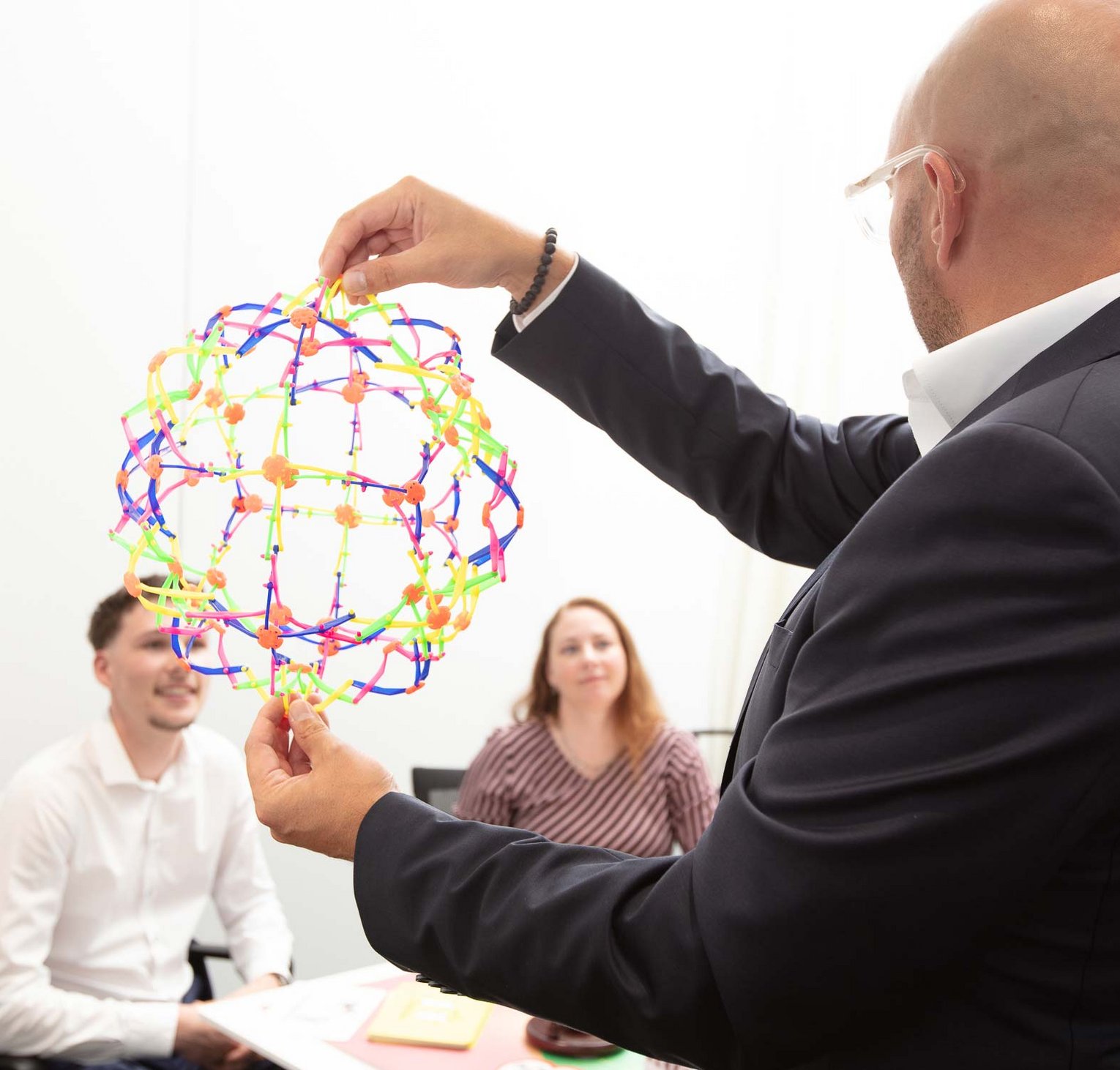 Sales seminars by Philipp Gotterbarm A man in a suit holds and demonstrates a colorful geometric sphere toy while two seated people attentively watch during a meeting or presentation.