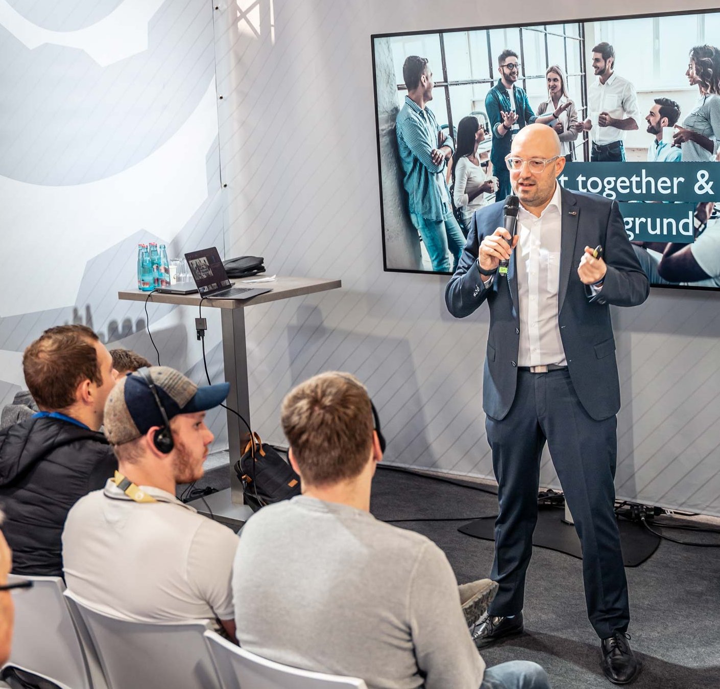 Sales training by Philipp Gotterbarm A man in a suit is giving a presentation to an audience seated in front of him. A large screen behind him displays an image of a group discussion with the words “together &” visible. The setting appears professional and focused on teamwork or collaboration.