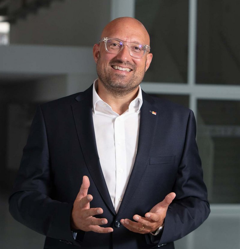 Philipp Gotterbarm: A Portrait A smiling bald man wearing glasses and a dark suit stands indoors, gesturing with his hands as if speaking or presenting.