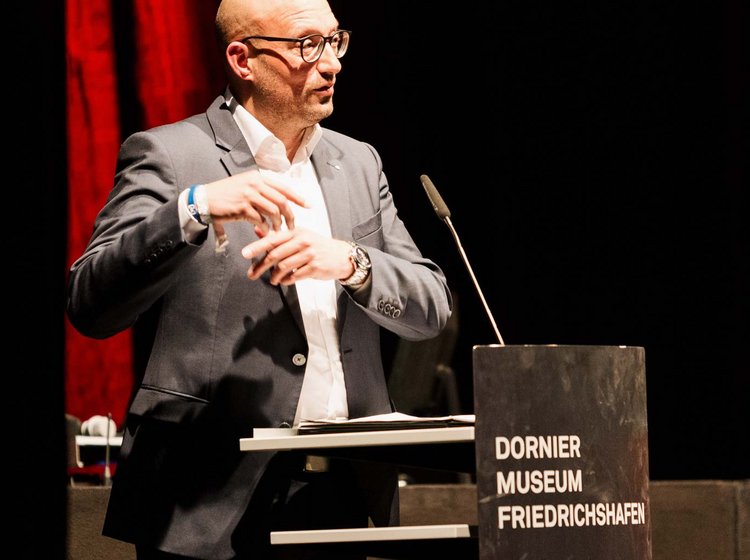 Philipp Gotterbarm at the lectern A man in a suit and glasses speaks at a podium labeled "Dornier Museum Friedrichshafen" during a presentation or lecture.