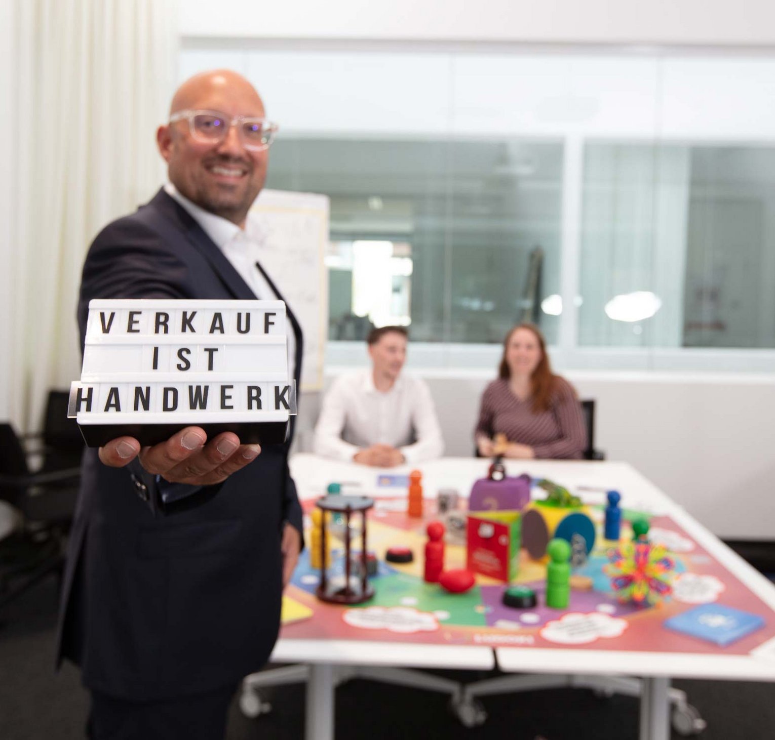 Sales training by Philipp Gotterbarm A smiling man in business attire holds a sign reading "VERKAUF IST HANDWERK" in a modern office, with two people sitting at a table covered in colorful toys and objects in the background.