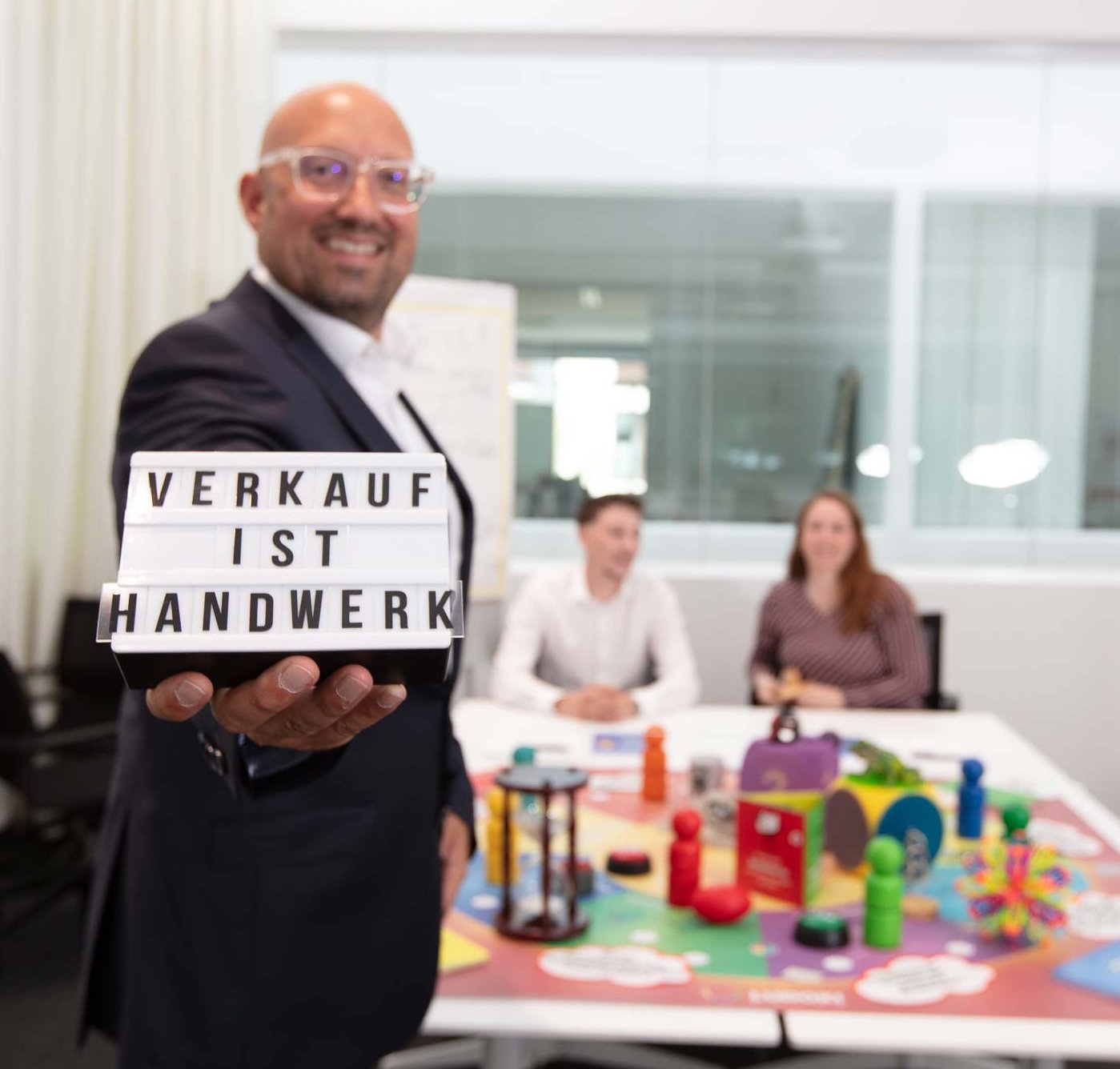 Sales training by Philipp Gotterbarm A smiling man in business attire holds a sign reading "VERKAUF IST HANDWERK" in a modern office, with two people sitting at a table covered in colorful toys and objects in the background.